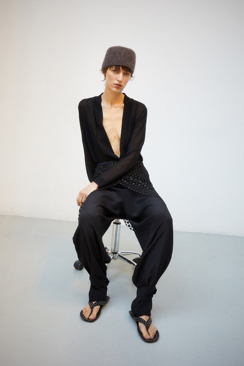 Vanherèen curated archive selection. Model seated wearing sheer black deep-v bodysuit and textured trousers. Made-to-order Italian luxury.